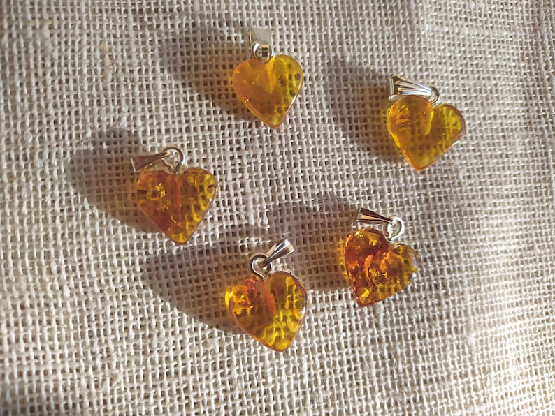 May include: Five amber heart-shaped pendants with silver bail loops. The pendants are arranged in a circle on a beige fabric background.