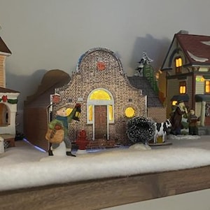 Może przedstawiać: Scena wioski bożonarodzeniowej z podświetlanymi miniaturowymi domami, figurkami i zaśnieżonym krajobrazem. Domy mają szczegółową architekturę i ciepłe oświetlenie wnętrz. Ekspozycja jest ustawiona na półce, tworząc uroczą świąteczną scenę.