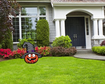 May include: A carved wooden pumpkin with a witch's hat and a silly face. The pumpkin is orange and the hat is black with a purple band. The pumpkin is in front of a house with a green lawn.