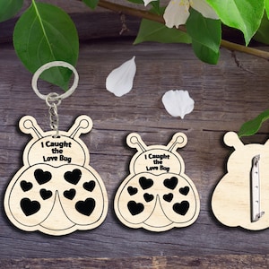 Può includere: Portachiavi in legno a forma di coccinella con il testo "I Caught the Love Bug" e cuori neri. Il portachiavi è mostrato con un anello e una catena di metallo.