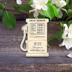 May include: Wooden gas pump ornament with the text "SELF SERVE" at the top. The pump displays funny fuel options and the year "2022" with the phrase "The year we couldn't afford Gas."