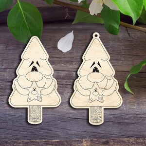 May include: Two wooden Christmas tree ornaments with a cartoon face holding a star. The ornaments are light brown with black outlines and details. The background is a dark wood surface with green leaves and white flower accents.