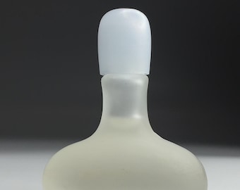 May include: A frosted glass perfume bottle with a pale blue cap. The bottle has a rounded base and a slender neck. The cap is a solid, rounded shape. The bottle is opaque and sits on a white surface.