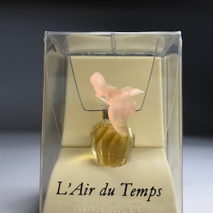 Op de afbeelding: Een klein, doorzichtig glazen flesje parfum met een roze vogelvormige dop. Het flesje zit in een witte doos met de tekst "L'Air du Temps Nina Ricci" op de voorkant.