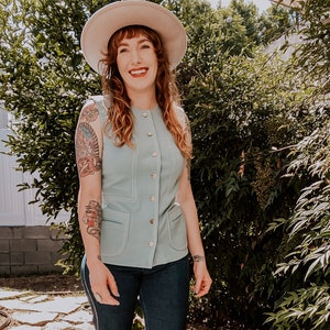 May include: A woman wearing a light blue sleeveless button-up vest with a white hat and dark blue jeans.