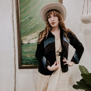 May include: A woman wearing a black velvet jacket with colorful patchwork details, a white hat, a white skirt with lace trim, and brown boots. She is standing in front of a painting.