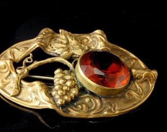 Op de afbeelding: Een vintage messing broche met een rode glazen steen en een druivenontwerp. De broche is in Art Nouveau-stijl.