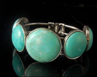 Op de afbeelding: Een zilveren armband met vijf turquoise cabochons. De stenen zijn gezet in zilveren bezels en zijn in een rij rond de armband gerangschikt.