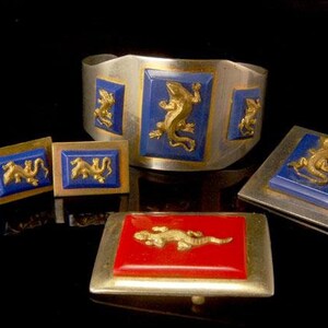 Puede incluir: Un conjunto de joyas de plata y oro con esmalte azul y rojo con un diseño de lagarto. El conjunto incluye un brazalete, dos pendientes y un broche.