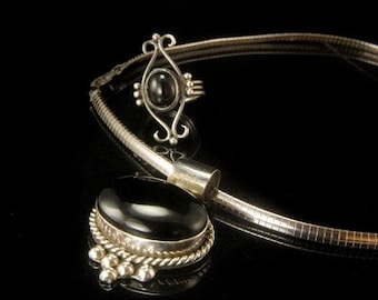 May include: A silver necklace with a black gemstone pendant and a matching ring. The necklace is a simple chain with a small, round pendant. The ring has a more elaborate design with a larger, oval-shaped gemstone.
