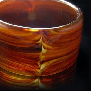 Puede incluir: Un objeto cilíndrico de vidrio con una paleta de colores cálidos de naranja, marrón y amarillo. El vidrio tiene un patrón arremolinado y en capas, y el borde es liso. El objeto se refleja en una superficie oscura.