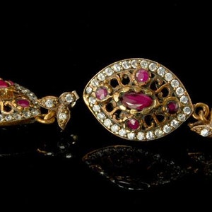 Może przedstawiać: Para ozdobnych, złocistych kolczyków z rubinami i diamentami. Każdy kolczyk ma centralny rubin otoczony mniejszymi rubinami i przezroczystymi, błyszczącymi diamentami. Projekt jest skomplikowany i szczegółowy, o estetyce vintage.