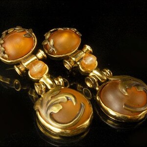 May include: A pair of gold tone earrings with amber colored glass stones. The earrings feature a floral design and are accented with smaller, pearlescent stones.
