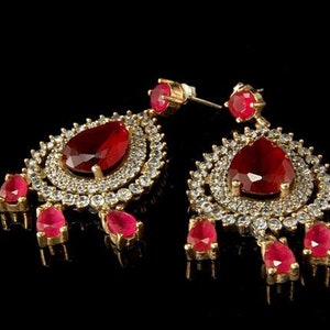 Może przedstawiać: Para złotych kolczyków z dużymi, czerwonymi kamieniami szlachetnymi w kształcie łezki. Kolczyki otoczone są aureolą z mniejszych, przezroczystych kamieni szlachetnych. Dodatkowe czerwone kamienie zwisają z dołu.