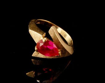 May include: A gold ring with a red gemstone set in a unique, abstract design.