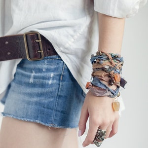 May include: A denim mini skirt with a frayed hem and a brown leather belt with a large buckle. A wrapped bracelet with fabric and beads adorns the wrist. A silver ring with multiple bands is on the finger.