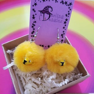 Puede incluir: Un par de pendientes amarillos y peludos con un diseño de abeja negra y amarilla. Los pendientes están empaquetados en una pequeña caja con el texto "Weasley's Wizard Wheezes" en la parte delantera.