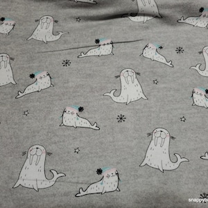 May include: A gray fabric with a repeating pattern of white walruses wearing blue and pink hats. The walruses are all facing forward with their mouths open and their tusks showing. The fabric is perfect for sewing into a baby blanket or other items for a nursery.