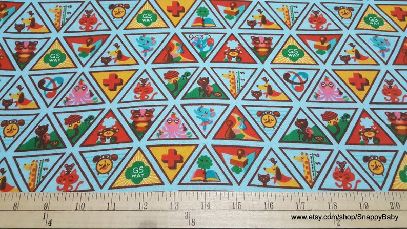 Puede incluir: Una tela azul con un patr&oacute;n repetitivo de parches triangulares coloridos. Cada parche presenta un dise&ntilde;o diferente de insignia de Girl Scout, incluyendo una cruz roja, un libro, un reloj, un gato, una jirafa y un pulpo. El texto "GS WAY" aparece en algunos de los parches.