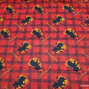 May include: Red and black checkered fabric with a repeating pattern of the Gryffindor house crest. The crest features a lion in a shield with the letter "G" on it.
