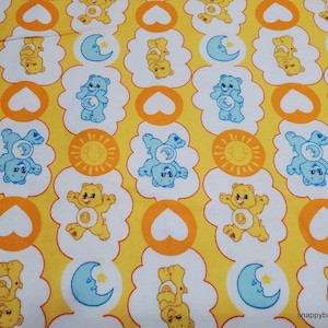 May include: Yellow fabric with a repeating pattern of Care Bears. The bears are wearing yellow and blue outfits and are holding a sun, moon, or heart. The background is yellow with white clouds.