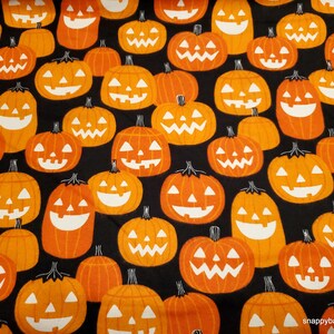 May include: A black fabric with a repeating pattern of orange jack-o'-lantern faces. The jack-o'-lanterns have white eyes, noses, and mouths.