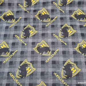 May include: Black and yellow fabric with a repeating pattern of a black badger inside a yellow shield with the text "HUFFLEPUFFTM" printed below the shield.