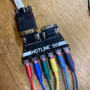 以下が含まれることがあります： 「HOTLINE S6」という文字が書かれた黒いデバイスに、6本の色のついたケーブルが接続されています。ケーブルの色は、赤、青、緑、黄、青、黒です。