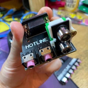 May include: A black circuit board with the text "HOTLINE" printed on it. The board has a variety of connectors, including a black VGA connector, two pink RCA connectors, and two black and silver connectors.