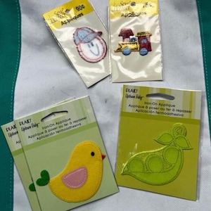 May include: Four iron-on appliques in their original packaging. The appliques include a smiling face wearing a baseball cap, a train, a yellow bird, and a green pea pod. The packaging includes the text "Sew Perfect Appliques" and "Uptown Baby".
