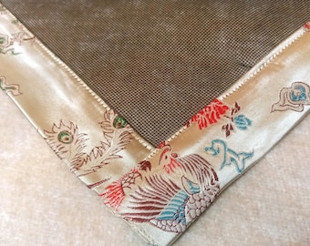 May include: A rectangular textile mat featuring a dark brown, fine-grid center panel bordered by a wide, cream-colored satin fabric. The border is decorated with intricate, multi-colored woven patterns of birds and floral motifs in red, blue, and gold.