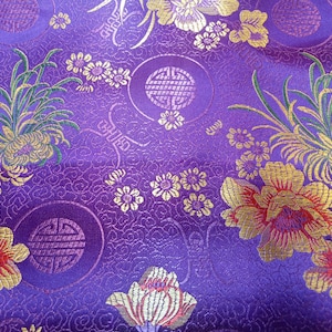 May include: Close-up of a vibrant purple fabric with an intricate floral pattern. Gold and red flowers, along with green foliage, are scattered across the surface. The design includes circular motifs and detailed textures, creating a rich, decorative textile.