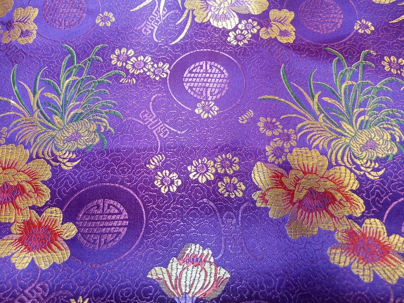 May include: Close-up of a vibrant purple fabric with an intricate floral pattern. Gold and red flowers, along with green foliage, are scattered across the surface. The design includes circular motifs and detailed textures, creating a rich, decorative textile.