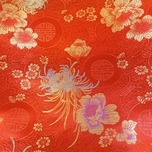 May include: Close-up of a vibrant red fabric with an intricate floral pattern. Gold and light blue flowers and foliage are scattered across the surface. The design includes circular motifs and small, detailed blossoms.