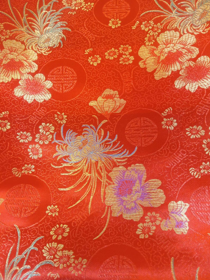 May include: Close-up of a vibrant red fabric with an intricate floral pattern. Gold and light blue flowers and foliage are scattered across the surface. The design includes circular motifs and small, detailed blossoms.