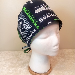 May include: A black and green surgical scrub hat with the Seattle Seahawks logo and team name repeated on the fabric. The hat has a tie on the back.