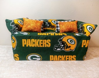Może przedstawiać: Miniaturowa sofa z zielono-złotą tkaniną Packers, z logo drużyny i napisem "Packers". Zawiera trzy ozdobne poduszki: dwie pomarańczowe we wzory i jedną w czarno-białą kratę.