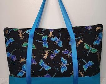 May include: A black tote bag featuring a repeating pattern of blue, green, and gold dragonflies. The bag has a solid teal blue base and two long, bright blue nylon straps. A zipper closure runs along the top edge of the rectangular fabric bag.