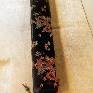 May include: A black rectangular case with a dragon design in red, gold, and green. The case has a red drawstring with green beads at the bottom. The dragon design is repeated along the length of the case.