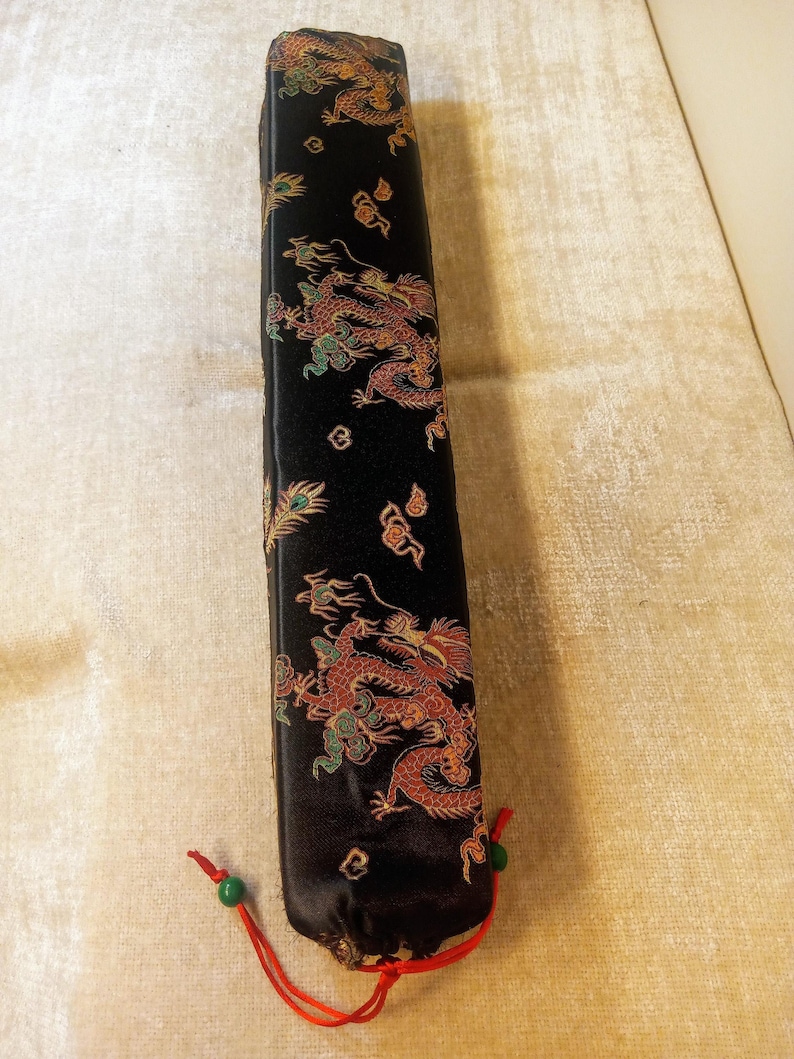 May include: A black rectangular case with a dragon design in red, gold, and green. The case has a red drawstring with green beads at the bottom. The dragon design is repeated along the length of the case.