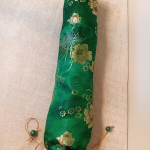 May include: A green, cylindrical fabric case with a floral pattern in gold and cream. The case has a drawstring closure with a green bead. The fabric has a sheen, and the design includes stylized flowers and foliage.