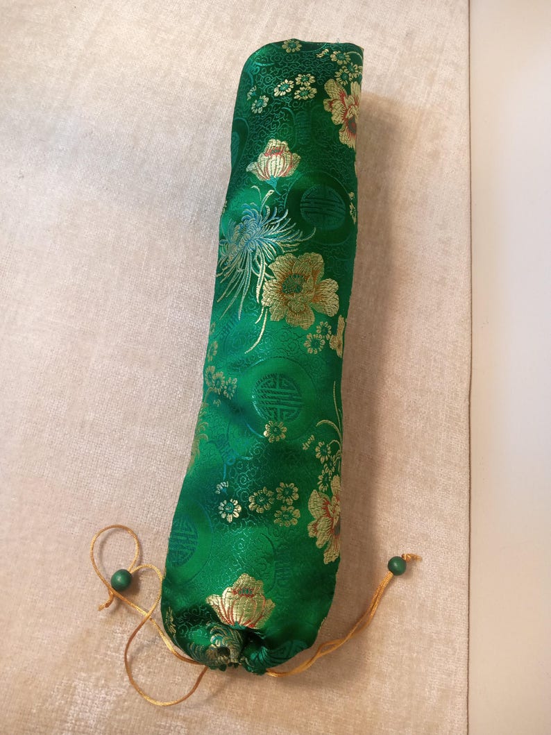 May include: A green, cylindrical fabric case with a floral pattern in gold and cream. The case has a drawstring closure with a green bead. The fabric has a sheen, and the design includes stylized flowers and foliage.