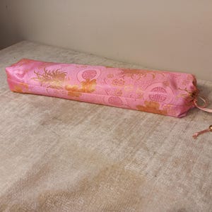 May include: A pink, rectangular, silk-like fabric case with a drawstring closure. The case features a gold floral and geometric pattern. The fabric is a vibrant pink with gold accents, creating a decorative and elegant appearance.