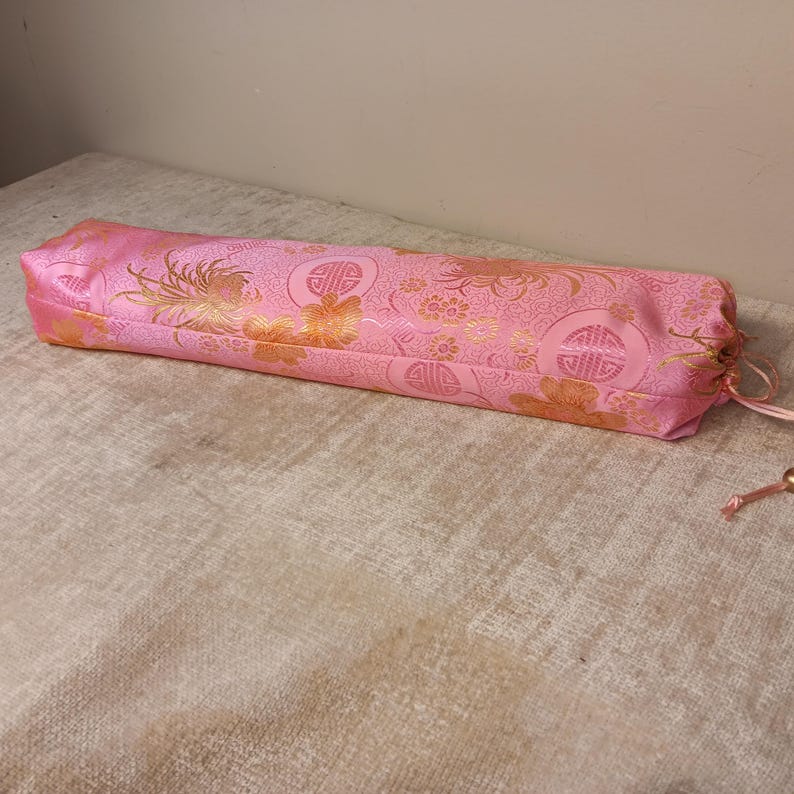 May include: A pink, rectangular, silk-like fabric case with a drawstring closure. The case features a gold floral and geometric pattern. The fabric is a vibrant pink with gold accents, creating a decorative and elegant appearance.