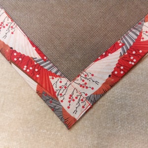 Może przedstawiać: Kwadratowy kawałek tkaniny z ciemnobrązowym siatkowym środkiem otoczonym materiałową lam&oacute;wką. Brzeg ozdobiony jest powtarzającym się wzorem czerwono-białych składanych wachlarzy przeplatanych czarnymi gałązkami i małymi czerwonymi kwiatami.