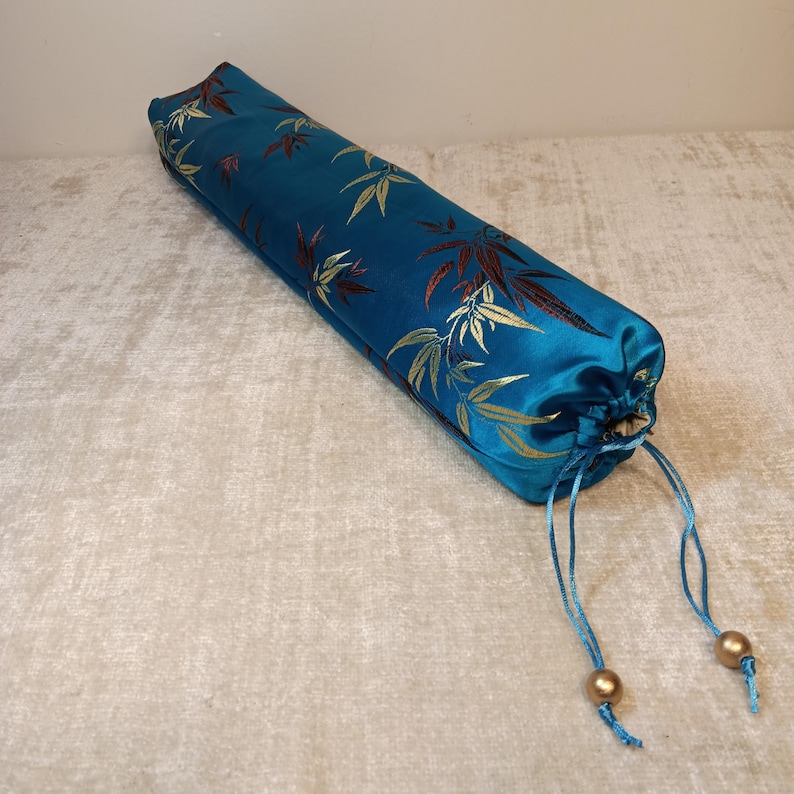 May include: A cylindrical teal-colored yoga bolster with a bamboo leaf pattern in gold and burgundy. The bolster has a drawstring closure with two gold-colored beads. The fabric has a satin sheen.