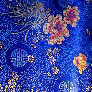May include: Close-up of a vibrant blue fabric with an intricate floral pattern. The design features large flowers in shades of gold, red, and pink, along with smaller yellow blossoms and circular motifs. The background is filled with swirling patterns.