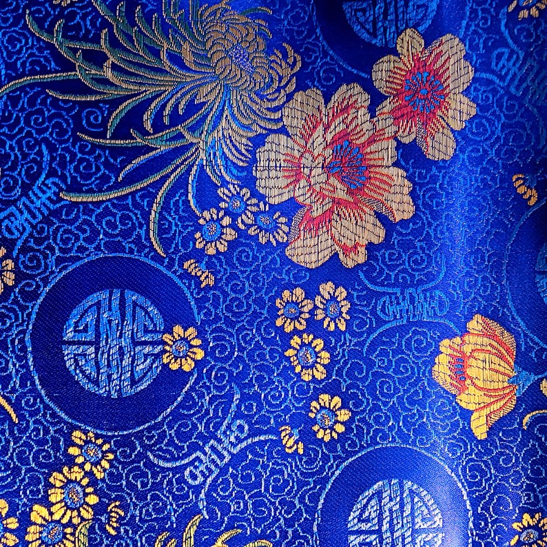 May include: Close-up of a vibrant blue fabric with an intricate floral pattern. The design features large flowers in shades of gold, red, and pink, along with smaller yellow blossoms and circular motifs. The background is filled with swirling patterns.