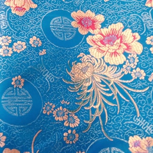 May include: A vibrant blue fabric with an intricate floral pattern. Gold and pink flowers and smaller blossoms are scattered across the surface. The design includes circular motifs with stylized symbols, creating a rich, decorative textile.