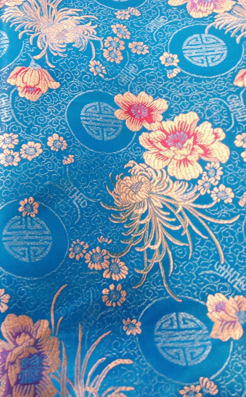 May include: A vibrant blue fabric with an intricate floral pattern. Gold and pink flowers and smaller blossoms are scattered across the surface. The design includes circular motifs with stylized symbols, creating a rich, decorative textile.